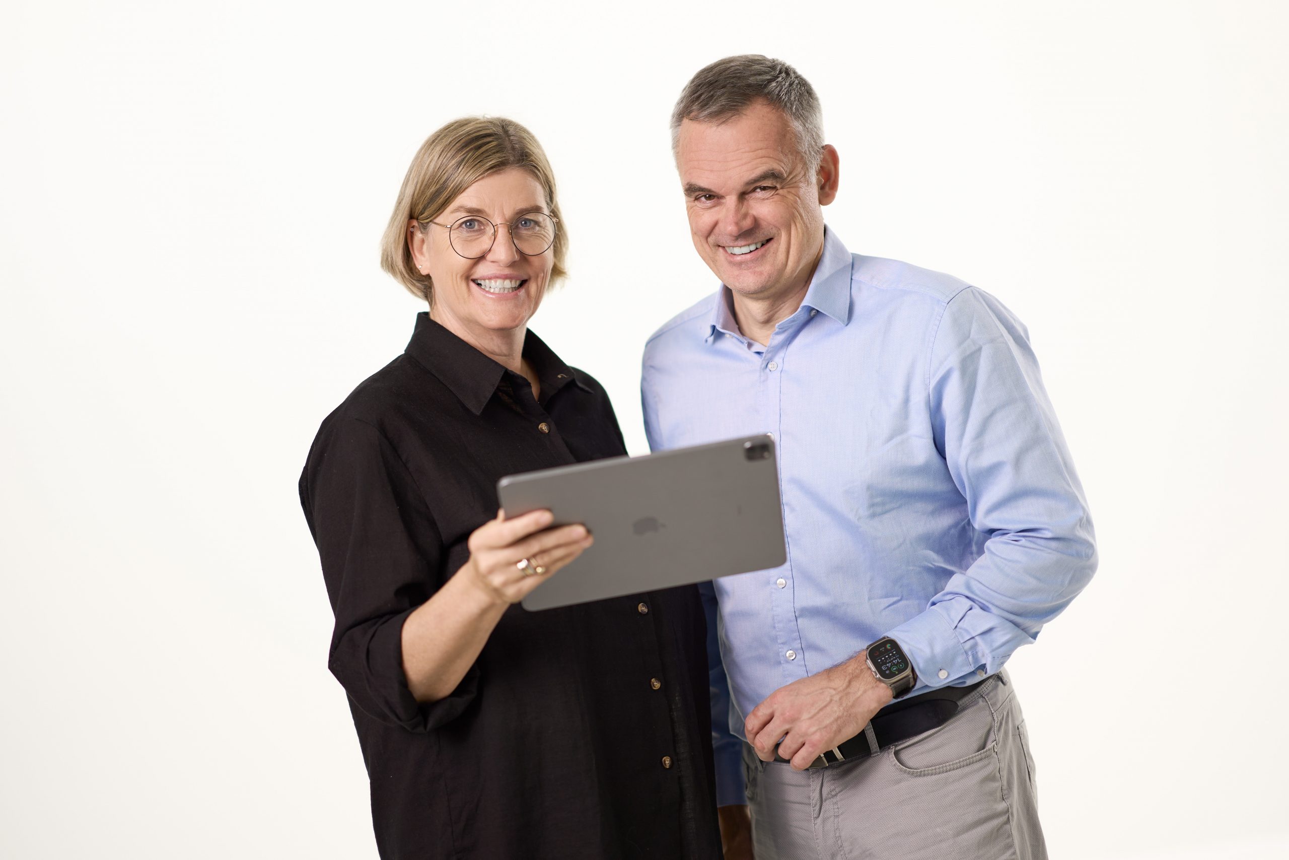 Stefani und Carsten stehen nebeneinander und schauen nach vorne. Sie halten ein iPad in der Hand und lächeln.