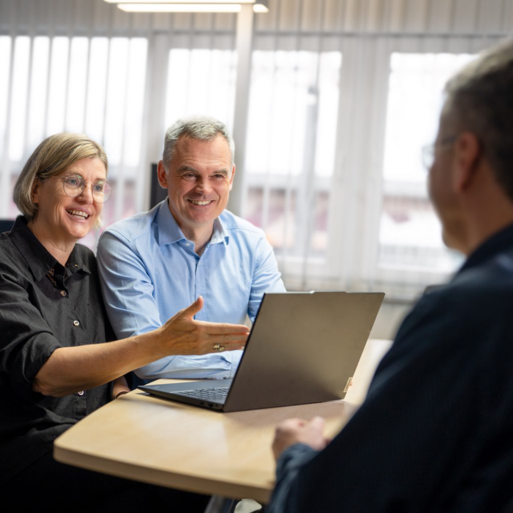 Stefani und Carsten sitzen nebeneinander an einem Schreibtisch. Sie sprechen mit einem Kunden.